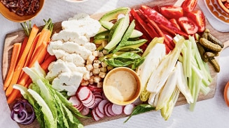 Bord met diverse gesneden groenten, zoals wortels, bloemkool, radijs, avocado, tomaat, rode paprika, bleekselderij, romaine sla, augurken en witlof, geserveerd met een kom dipsaus.