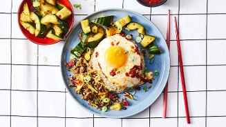 Gebakken rijstschotel met een spiegelei, komkommer en rode peper, geserveerd op een blauw bord met rode eetstokjes.