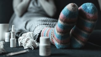 Persoon met wollen sokken, omringd door gebruikte papieren zakdoekjes en medicijnen