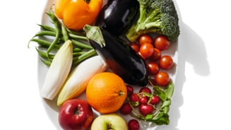 Assortiment van verse groenten en fruit op een witte schaal, inclusief paprika's, aubergines, broccoli, cherrytomaten, sperziebonen, witlof, sinaasappel, appels en radijsjes.
