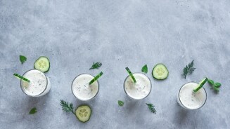 Vier glazen komkommer-yoghurtdrank op een grijze ondergrond, gegarneerd met schijfjes komkommer en kruiden.