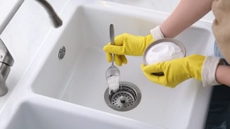 Persoon met gele rubberen handschoenen die poedervormig schoonmaakmiddel in een gootsteen schept