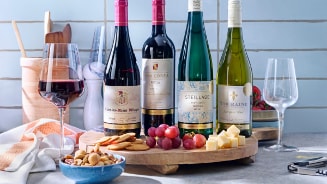 Vier flessen wijn op een houten plateau met bijpassende glazen, omringd door druiven, kaasblokjes, crackers en noten.