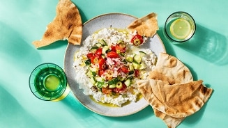 Mediterrane gerecht met geitenkaas, tomaten, en komkommer geserveerd met flatbread en een glas citroenwater.