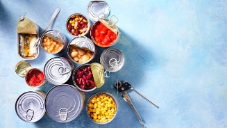 Opengewerkte blikken met verschillende soorten groenten en peulvruchten, zoals maïs, kidneybonen, kikkererwten, en tomatensaus, op een blauwe ondergrond met een blikopener erbij