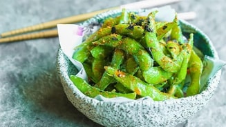 Bord met gekruide edamame bonen en eetstokjes op de achtergrond.