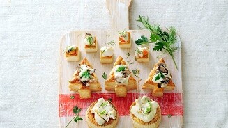 Verschillende feestelijke hapjes op een houten serveerplank, waaronder kerstboomvormige bladerdeeghapjes en ronde toastjes.