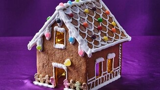 Peperkoekhuisje versierd met kleurrijke snoepjes en glazuur, op een paarse achtergrond.