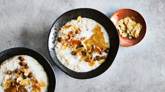 Havermoutpap met melk, geserveerd in zwarte kommen, gegarneerd met amandelschilfers, kaneel en een lepel fruitcompote, naast een schaaltje extra amandelschilfers.