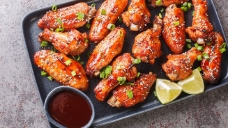 Knapperige kippenvleugels met saus, sesamzaad en lente-ui op een zwarte schaal, geserveerd met partjes limoen