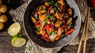 Kip cashew schotel met stukjes kip, paprika, ui, wortel en cashewnoten in een zwarte kom, geserveerd met limoen en eetstokjes