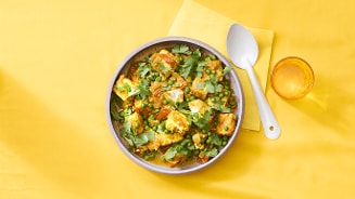 Schaal met Indiase curry van paneer, doperwten en koriander op een gele ondergrond, met lepel en glas.