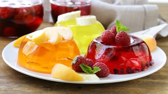 Drie kleurrijke gelatinepuddingen met vers fruit zoals perziken, bananen en frambozen op een wit bord.