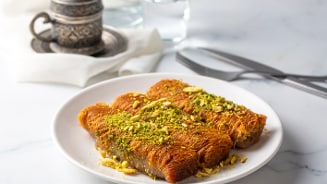 Twee stukken baklava met pistachenoten op een witte bord, met bestek en een kopje koffie op de achtergrond.
