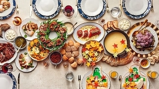 Feestelijke kersttafel vol gerechten, waaronder vlees, broodrolletjes, gepresenteerde kaasfondue, groentenkrans en diverse broodjes, omringd door gedekte borden en glazen.