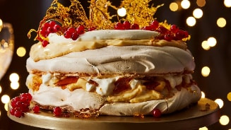 Pavlova taart gedecoreerd met rode bessen en gouden karamel op een taartstandaard, tegen een achtergrond van feestelijke lichtjes.