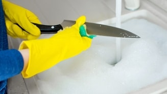 Afwassen van een keukenmes met gele rubberen handschoenen in een sopje bij een gootsteen.