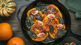 Pompoenpannenkoeken in een koekenpan, gegarneerd met verse vijgen, pecannoten en salieblaadjes, omringd door kleine pompoenen op een houten tafel