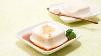 Zijden tofu geserveerd op een vierkant bordje met sojasaus en garneerblaadje munt, op de achtergrond nog een portie tofu met een houten lepel.