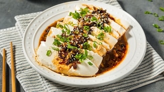 Plakjes zachte tofu geserveerd op een bord met pittige sojasaus, gegarneerd met gesneden bosui, sesamzaad en knapperige knoflook.