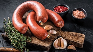 Gerookte worsten op een houten snijplank met knoflook, tijm, roze peperkorrels en grof zout op de achtergrond.