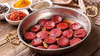 Gebakken plakjes sucuk in een koperen pan met specerijen op de achtergrond.