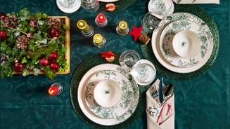 Feestelijk gedekte tafel met kerstdecoratie, groene tafelloper, porseleinen borden en brandende kaarsen.