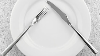 Wit bord met vork en mes geplaatst in een ruststand op een grijze tafel.