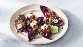 Gevulde paarse zoete aardappel met een topping van gehakt, crème fraîche, rode ui, rode peper en verse kruiden, geserveerd op een ovale schaal met een halve limoen.