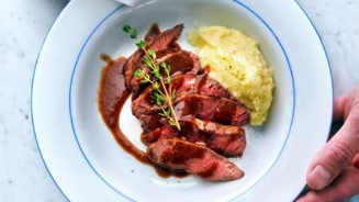 Bord met gesneden biefstuk en aardappelpuree, gegarneerd met een takje tijm, vastgehouden door een persoon.