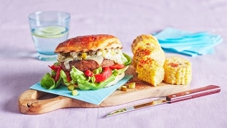 Hamburger met sla, tomaat en augurk op een houten plank, geserveerd met gegrilde maiskolven en een glas water met citroen.