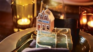 Tafeldecoratie met Allerhande miniatuur-huisje en cadeauverpakking, omgeven door kaarslicht.