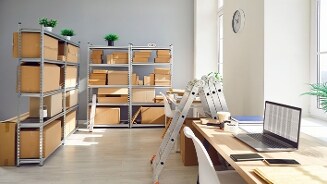 Magazijnruimte met metalen stellingen vol kartonnen dozen, bureaus met laptops en kantoorbenodigdheden, gevouwen ladders en veel daglicht door grote ramen.
