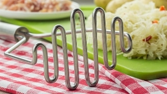 Roestvrijstalen pureestamper op een roodwit geruite doek met gesneden groenten op een groene snijplank op de achtergrond