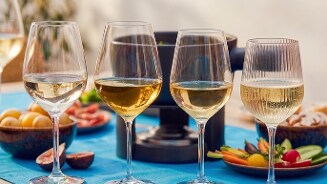 Vier glazen witte wijn op een blauwe tafel, omringd door schalen met vers fruit en groenten, met een fonduepan op de achtergrond.