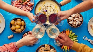 Vier mensen proosten met glazen wijn rond een tafel met kaasfondue, verschillende groentes, brood, aardappelen, champignons en vleeswaren.