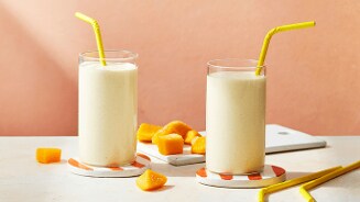 Twee glazen mango smoothie met gele rietjes, op onderzetters met oranje strepen, omringd door stukjes mango.