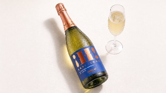 Fles mousserende wijn met blauwe Barcelona-label en een gevuld glas cava ernaast op een lichte ondergrond.