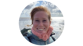 Persoon aan zee met een blauwe jas en roze sjaal op een zonnige dag, gezicht is wazig om privacy te beschermen.