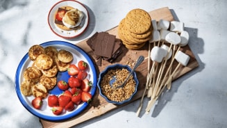 Poffertjes en aardbeien op een bord, met ingrediënten voor s'mores zoals chocolade, marshmallows op stokjes, koekjes en gehakte nootjes op een houten plank.