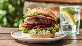 Vegetarische burger met sla, rode ui, augurk, bieten en een broodje met sesamzaad, geserveerd op een bord naast een glas water met citroen en munt.