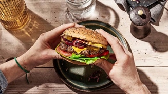 Een persoon houdt een hamburger vast met sla, tomaat, augurk, kaas en rode ui op een houten tafel.