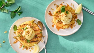 Twee borden met gegrilde ananasschijven, een bol vanille-ijs, gehakte pistachenoten, muntblaadjes en een partje citroen, geserveerd met karamelsaus.