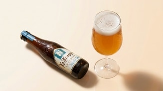Een flesje La Trappe Nillis 0.0% trappistenbier naast een gevuld bierglas op een lichtgekleurde achtergrond.