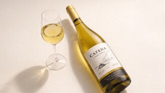 Fles Catena Chardonnay witte wijn naast een glas ingeschonken Chardonnay wijn op een lichte ondergrond.