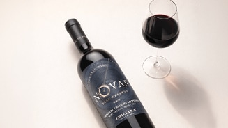 Fles rode wijn Novas Gran Reserva van het merk Emiliana, naast een glas rode wijn op een lichte ondergrond.