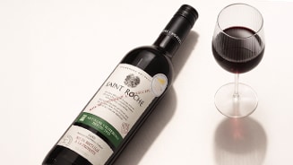 Fles rode wijn Saint Roche naast een gevuld wijnglas op een lichte ondergrond.