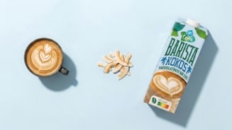 Kopje koffie met latte art, kokossnippers, koffiemeta en een pak Albert Heijn Terra Barista Haver Karamelsmaak op een lichtblauwe achtergrond.
