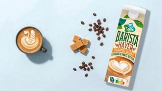 Kopje koffie met latte art, karamelblokjes, koffiemeta en een pak Albert Heijn Terra Barista Haver Karamelsmaak op een lichtblauwe achtergrond.