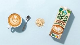 Kopje cappuccino met latte art, een lepel, een hoopje haverkorrels en een pak AH Terra Barista Haver plantaardige drank op een lichtblauwe ondergrond.